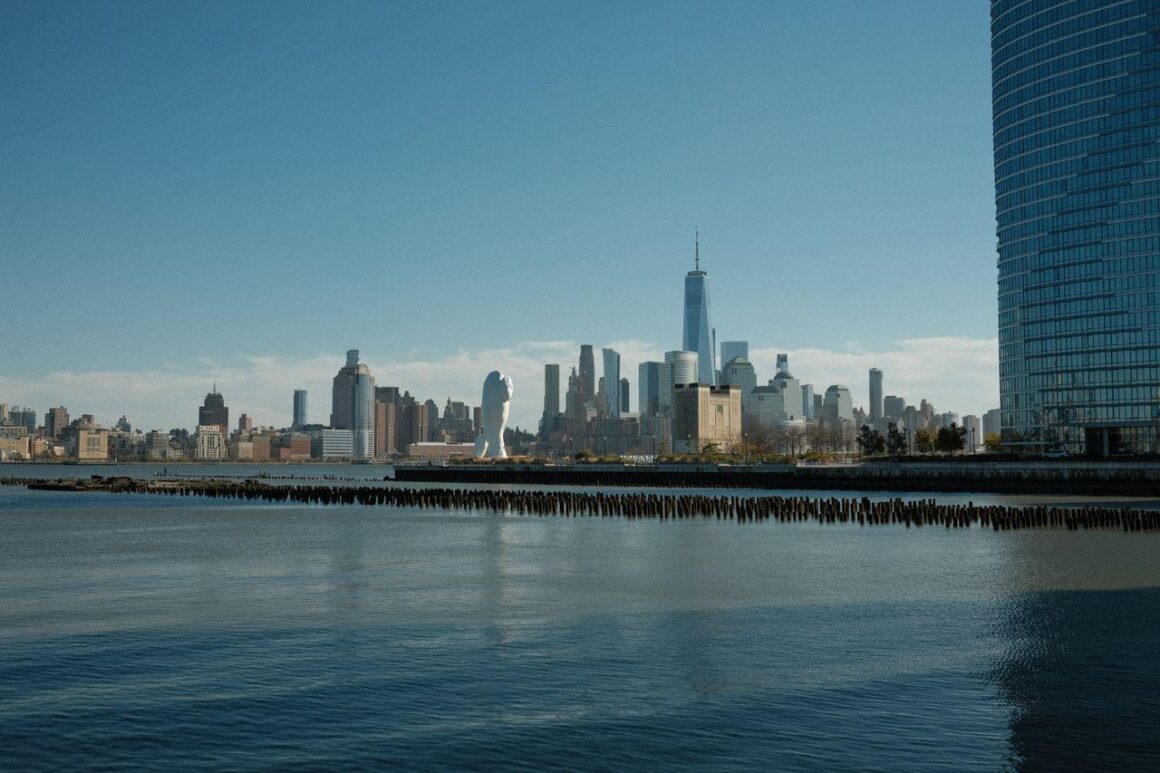 New York City skyline seen across the Hudson River from the Jersey City waterfront, with One World Trade Center rising above the surrounding Manhattan skyscrapers under a clear blue sky.